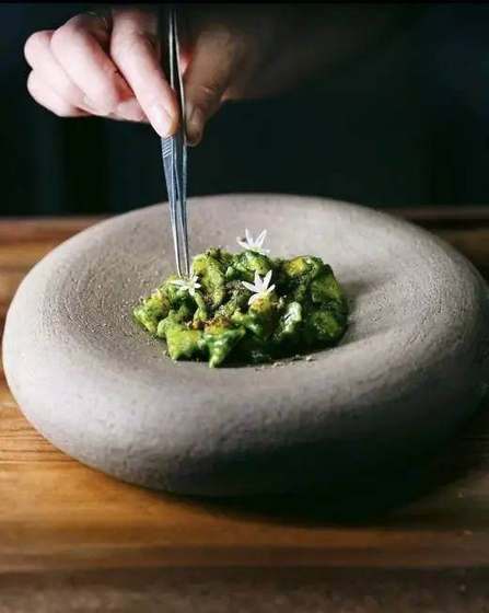 Un piatto di Indiniò: con una pinzetta si posano fiori bianchi su una pasta verde, in una ciotola di pietra